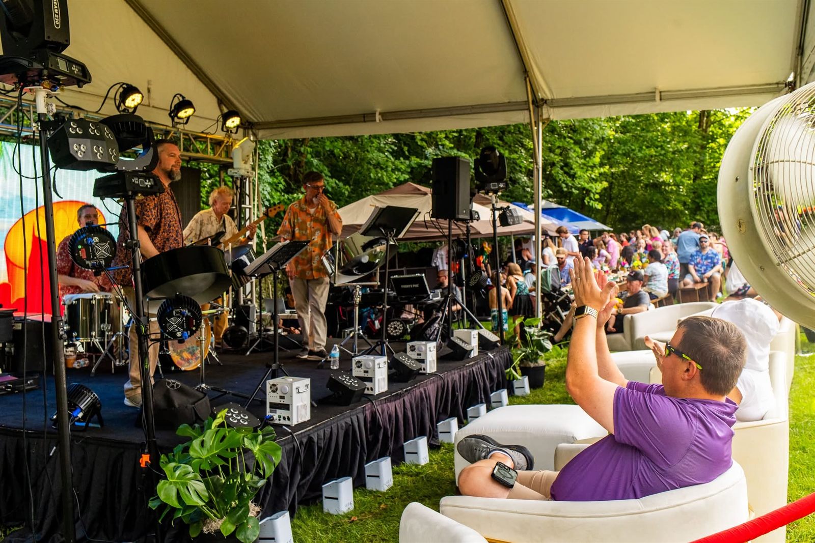 A live band performing on a skirted stage with lighting rigs under a tent while a guest applauds from a white lounge chair in a roped VIP section