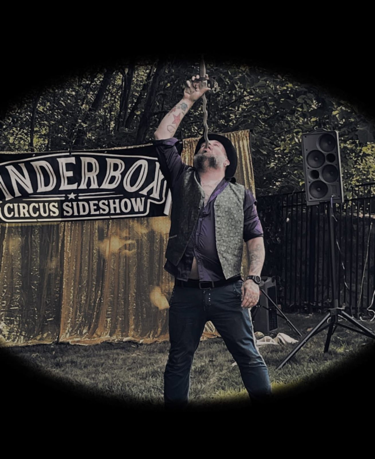 A sideshow performer in a bowler hat and vest swallowing a sword on a backyard stage in front of a gold curtain