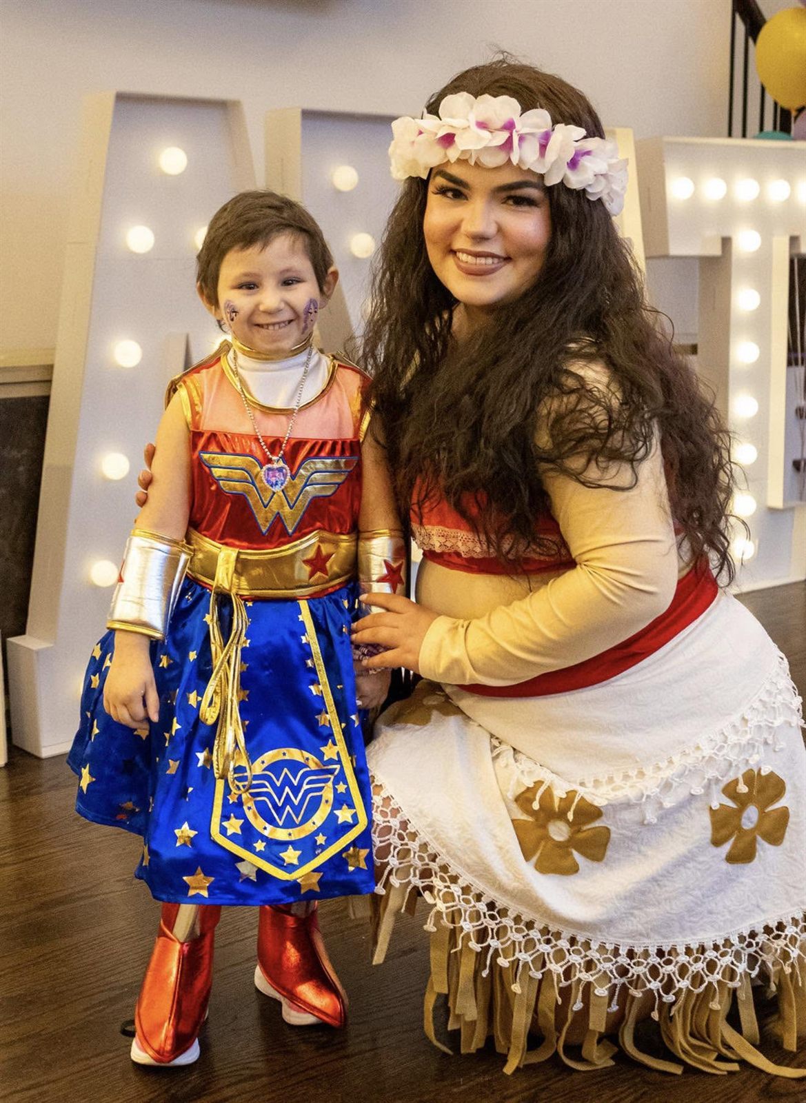 Island-princess character kneeling beside a child in a superhero costume by light-up marquee letters