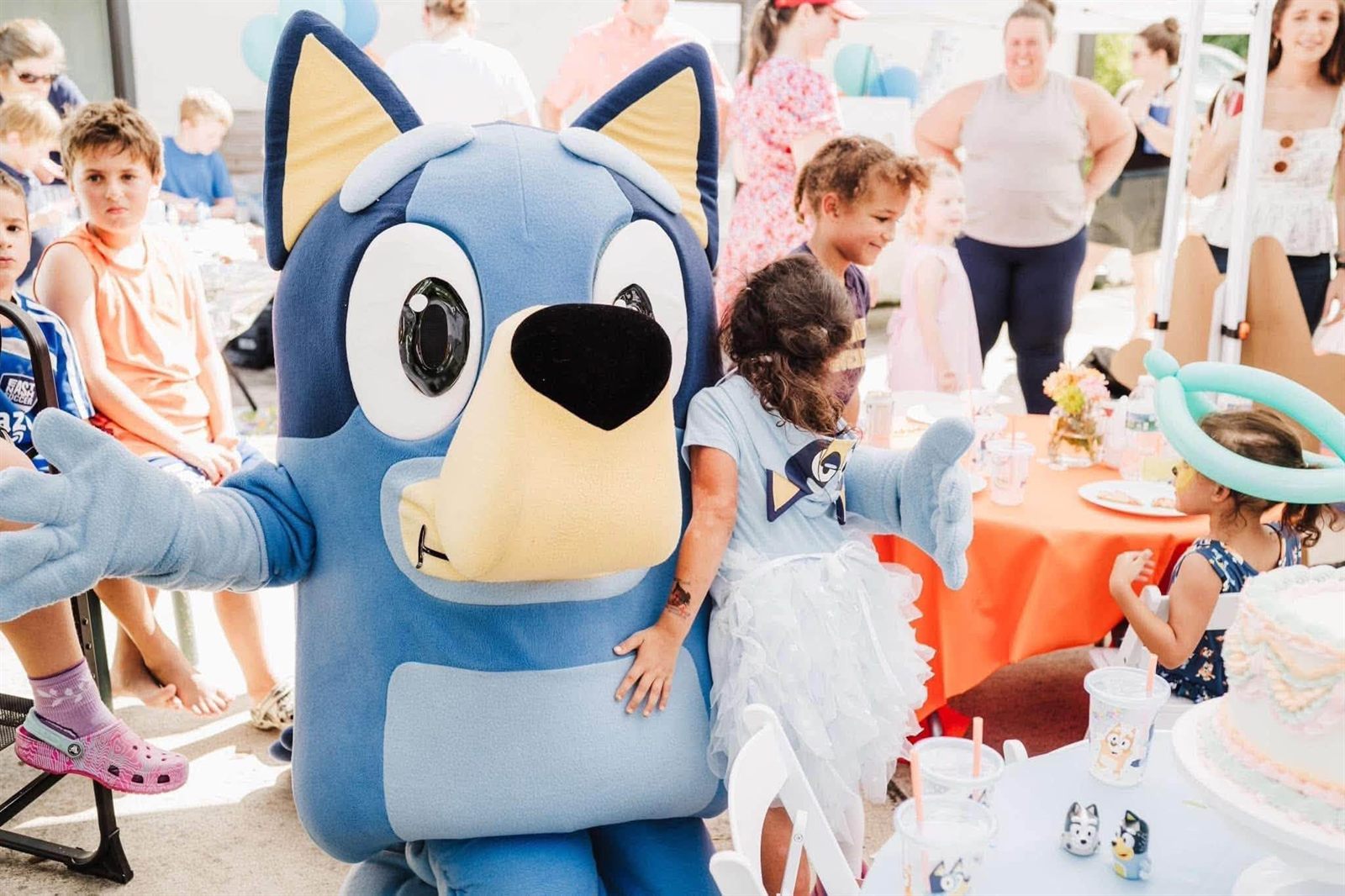A blue cartoon-dog mascot performer hugging a child among party guests at an outdoor birthday