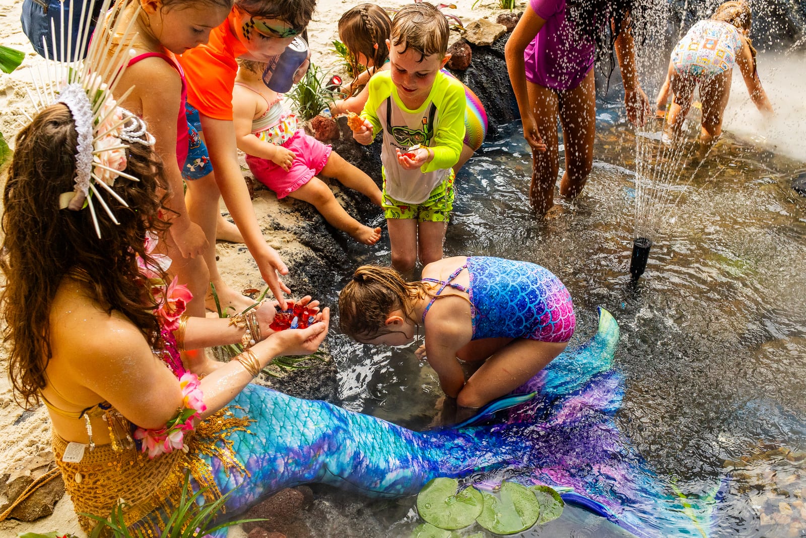 Mermaid performer in a blue sequinned tail seated beside a stream as children play in the water