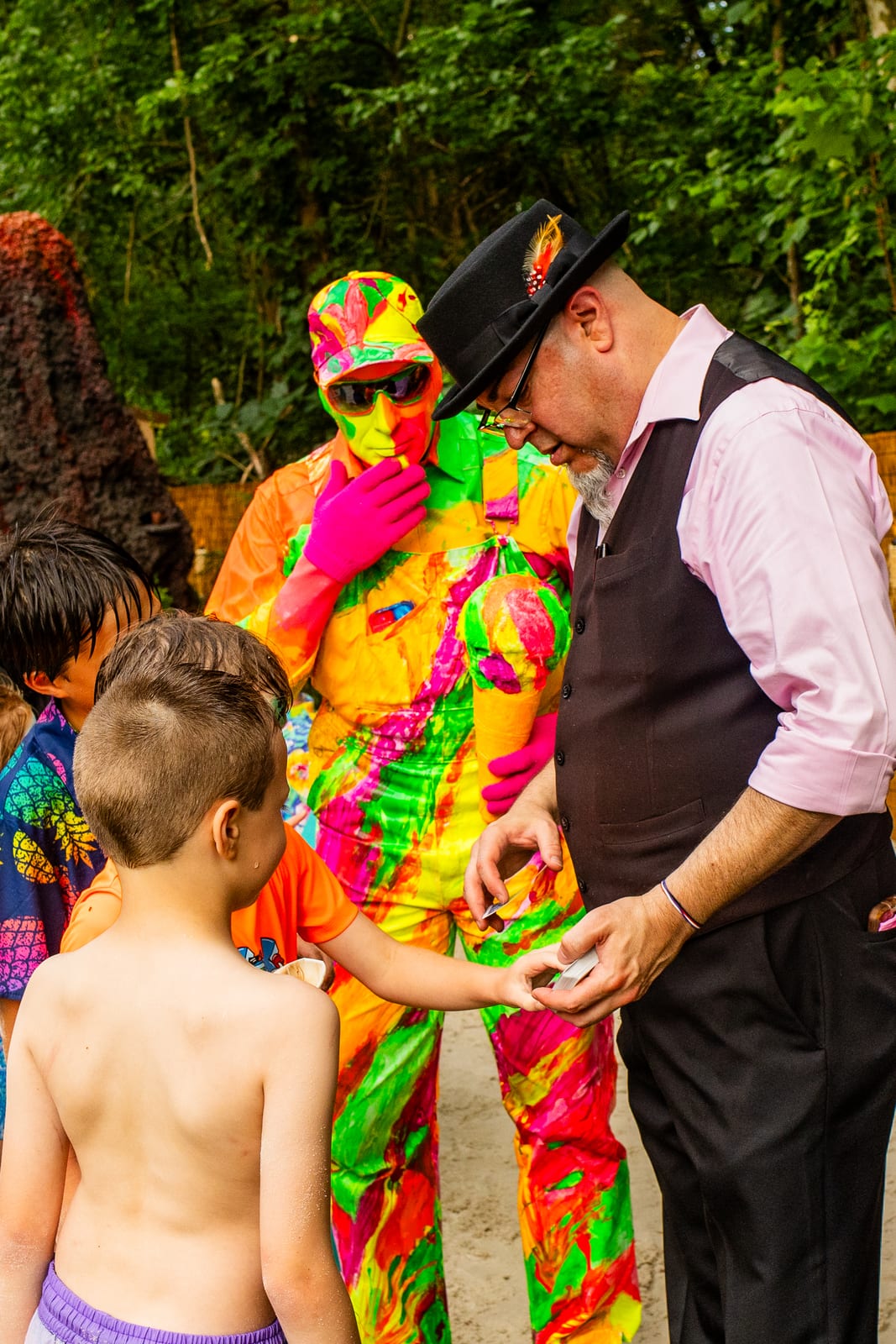 Magician performing close-up magic for a young guest beside a neon-costumed performer