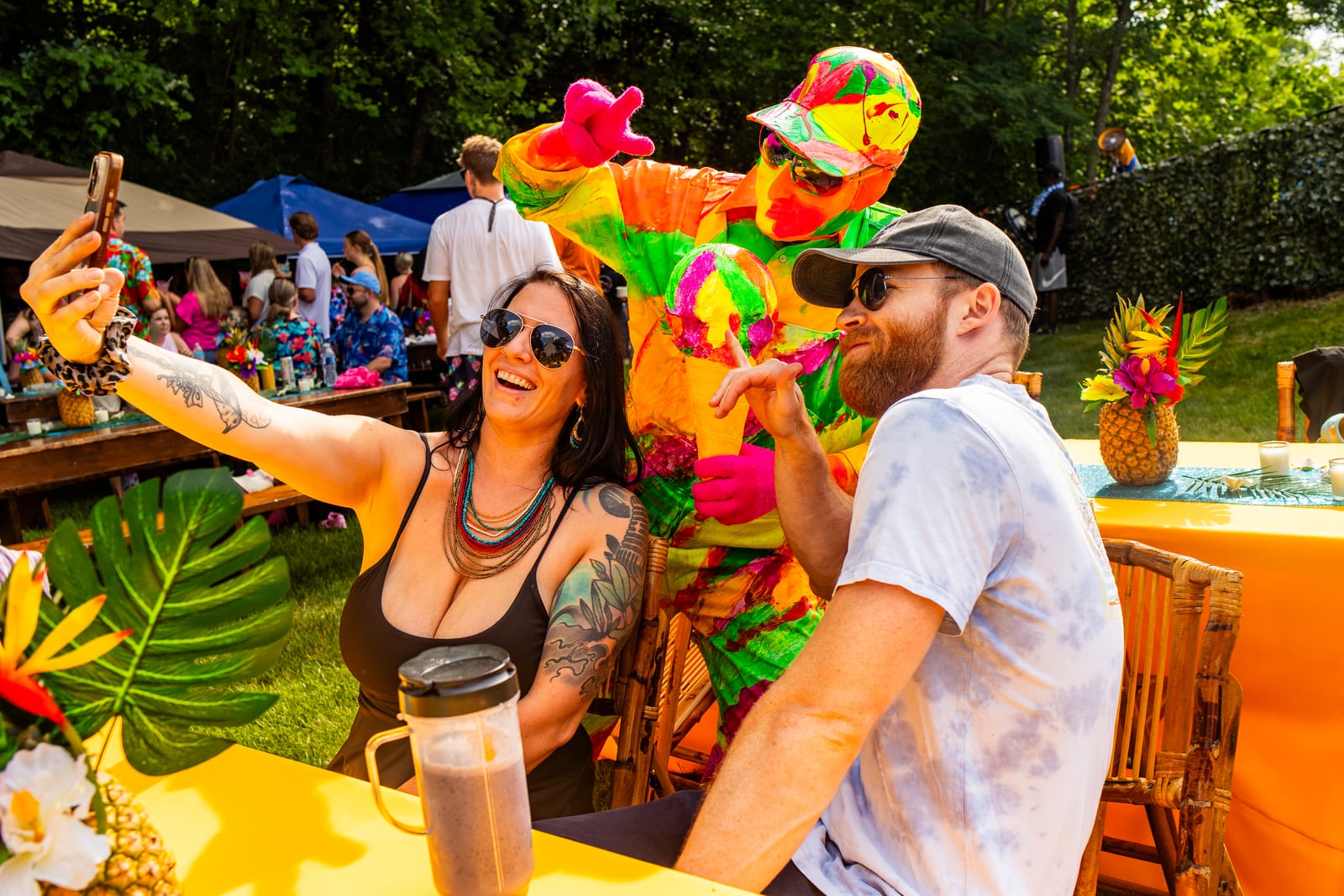 Guests taking a selfie at a tropical table as a neon-costumed performer leans in behind