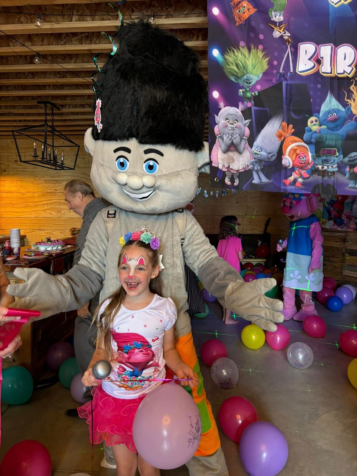 Blue-haired troll mascot dancing with a young guest among balloons at an indoor party