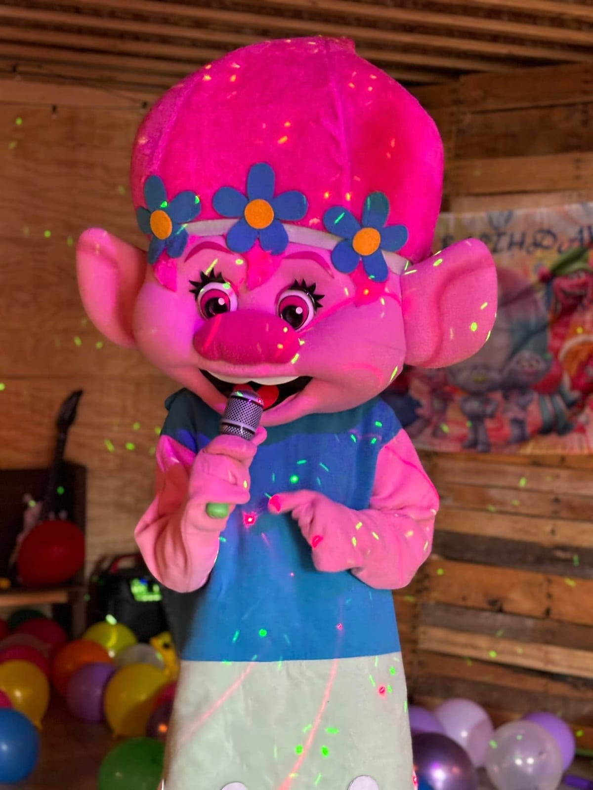 Pink-haired troll mascot with flower crown posing beside balloon decor