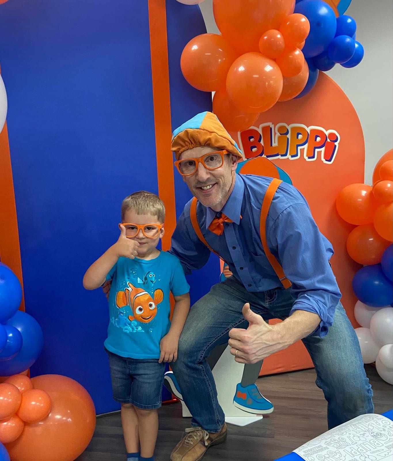 Orange-and-blue costumed children’s entertainer posing with a young guest by a balloon wall