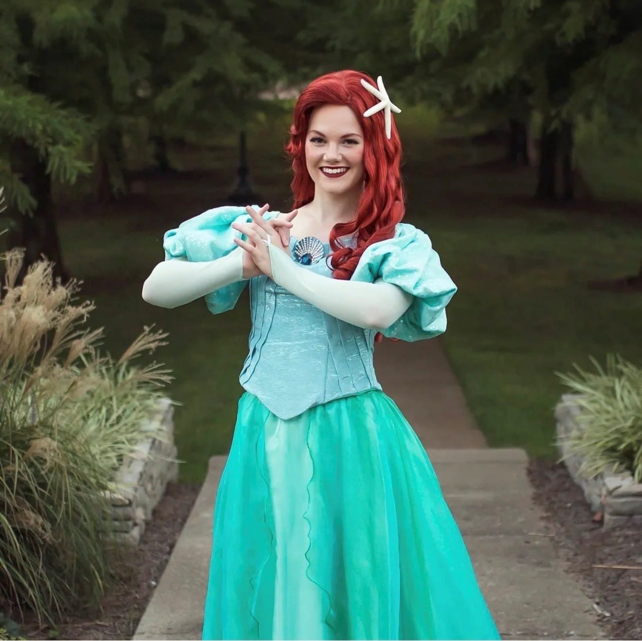 Red-haired princess character in an aqua and teal gown standing on a garden path