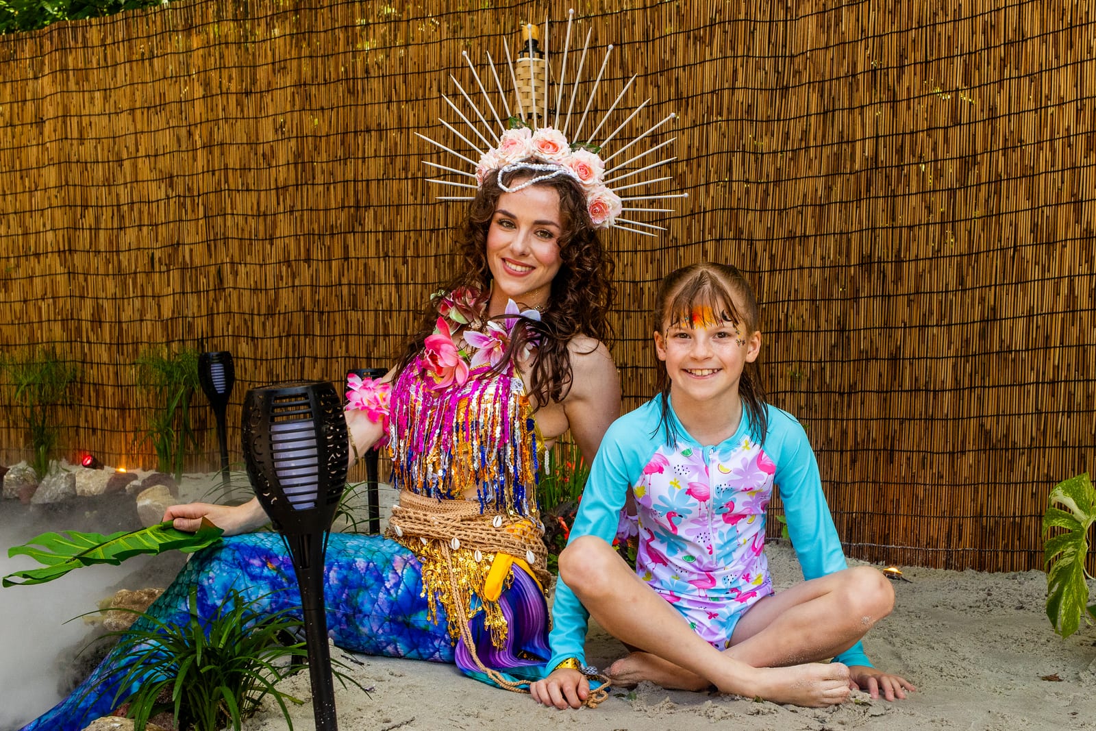 Mermaid performer in a sequinned tail seated on sand with a young guest beside tiki torches