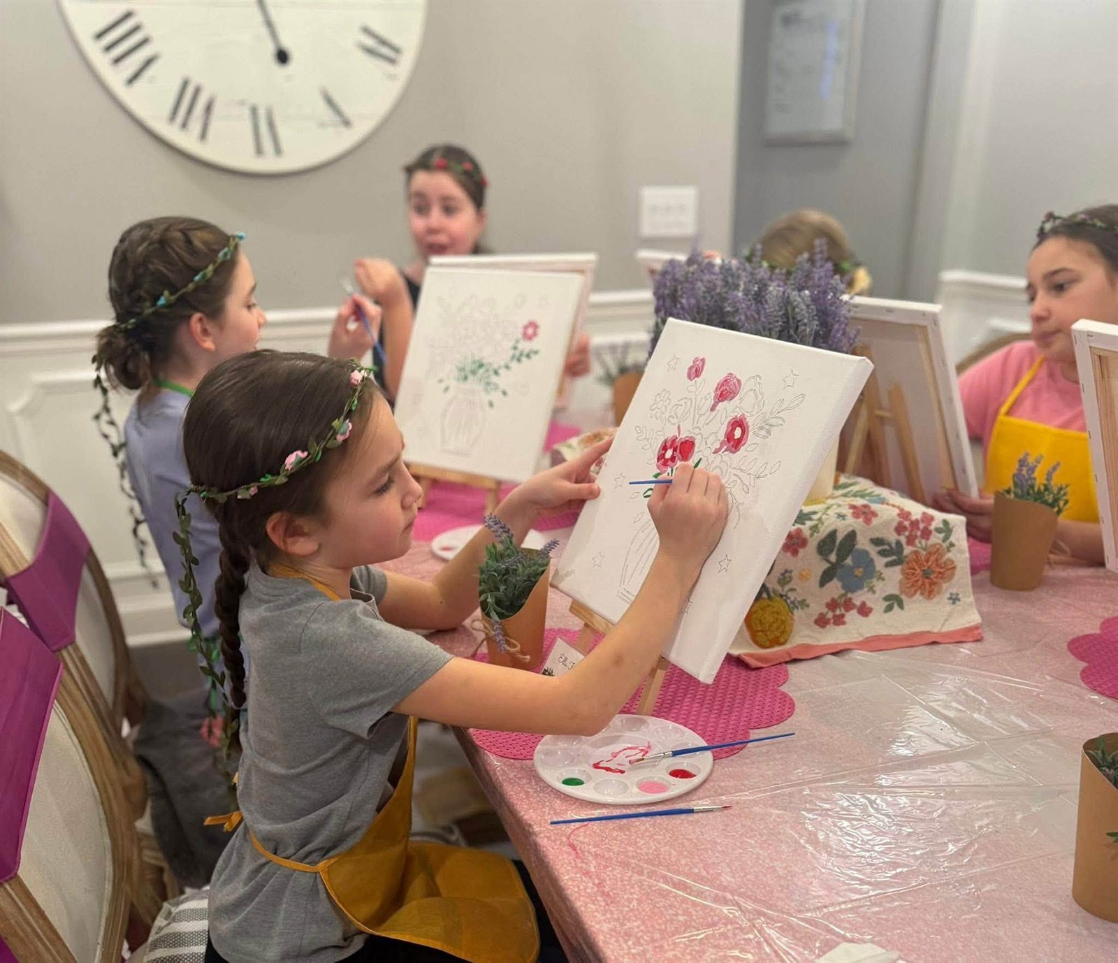 Children in flower crowns and aprons painting floral canvases at a pink-draped craft party table