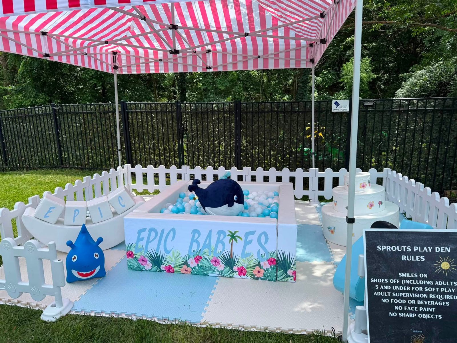 Toddler soft-play ball pit with a white picket fence, striped canopy and playful signage in a yard