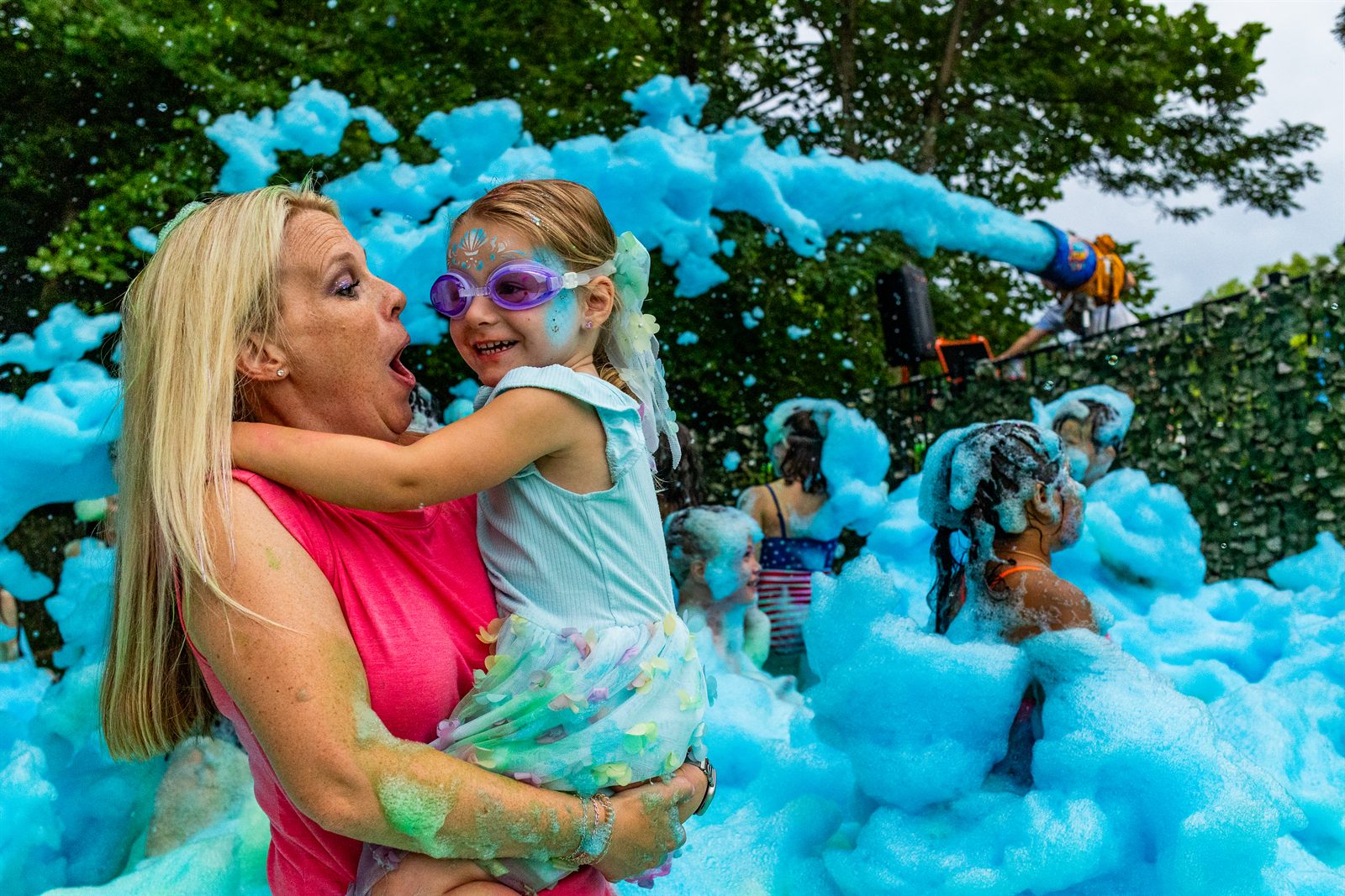 An adult and a goggled child laugh together surrounded by blue foam as guests play behind them