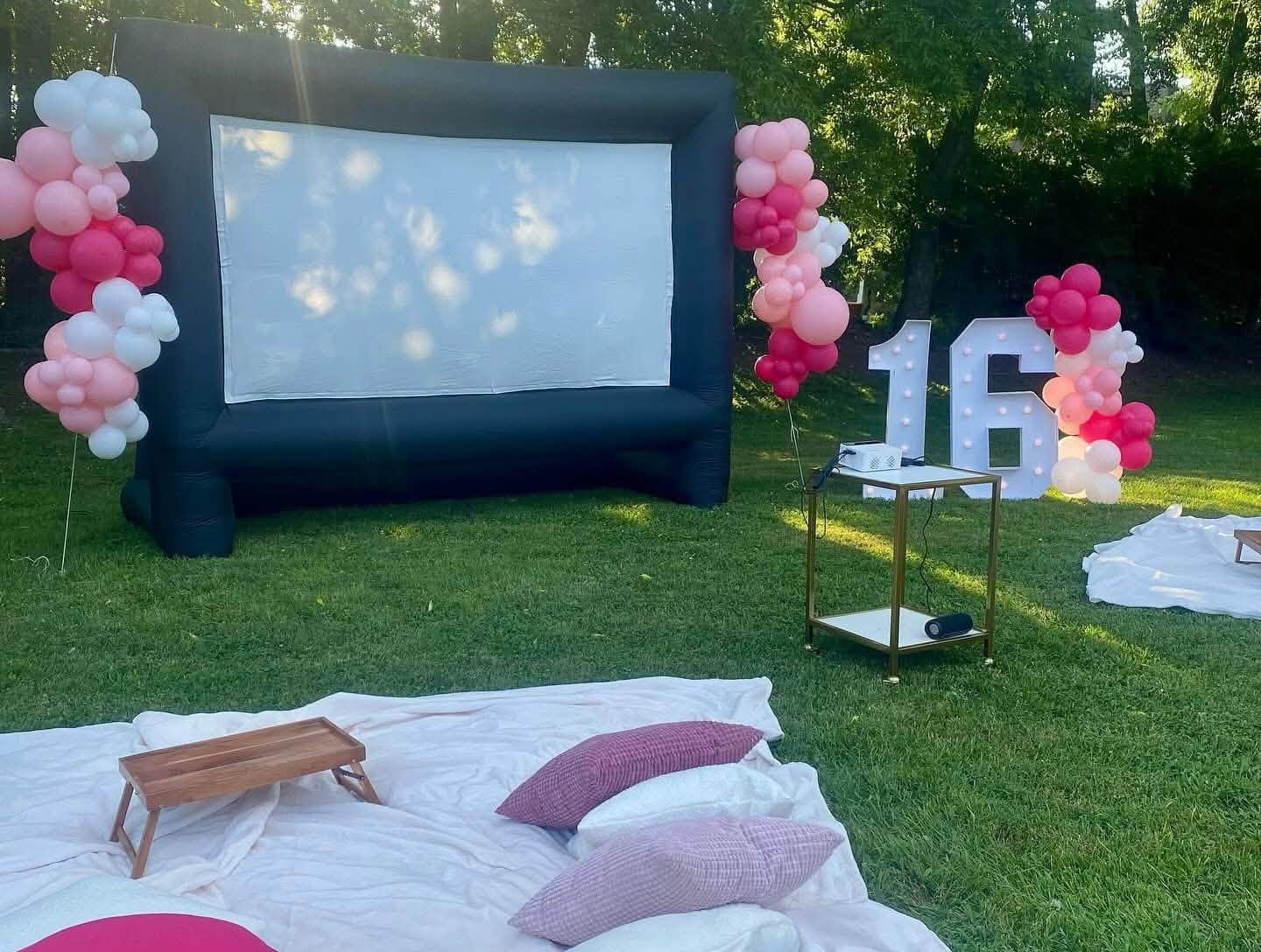 Inflatable movie screen framed by pink balloon garlands beside a lit 16 marquee on a lawn