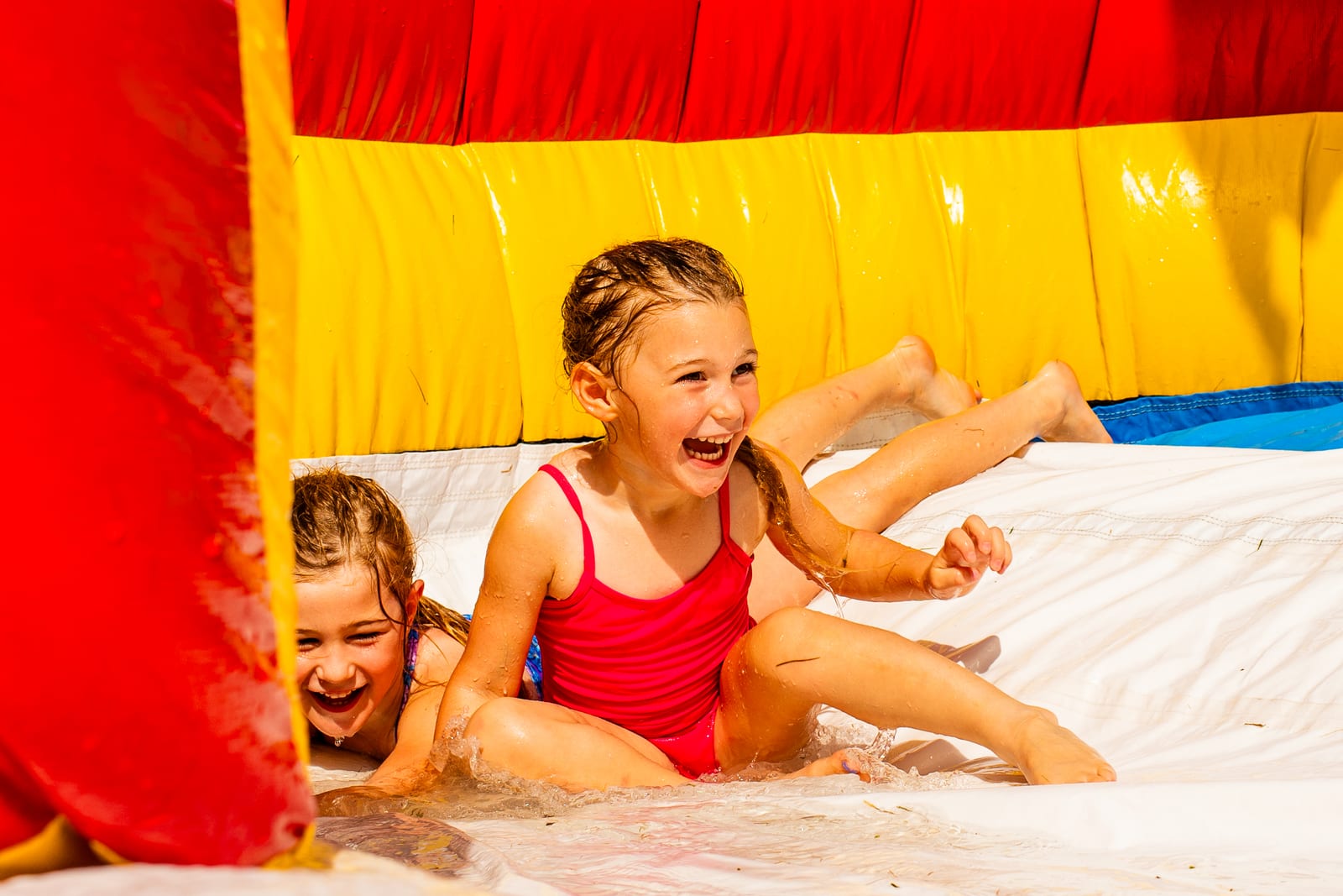 Two children sliding down an inflatable water slide together, both laughing