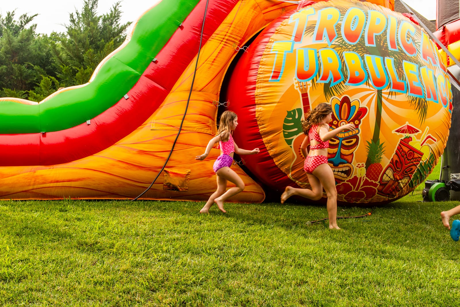 Two children running past a giant Tropical Turbulence inflatable slide on a lawn