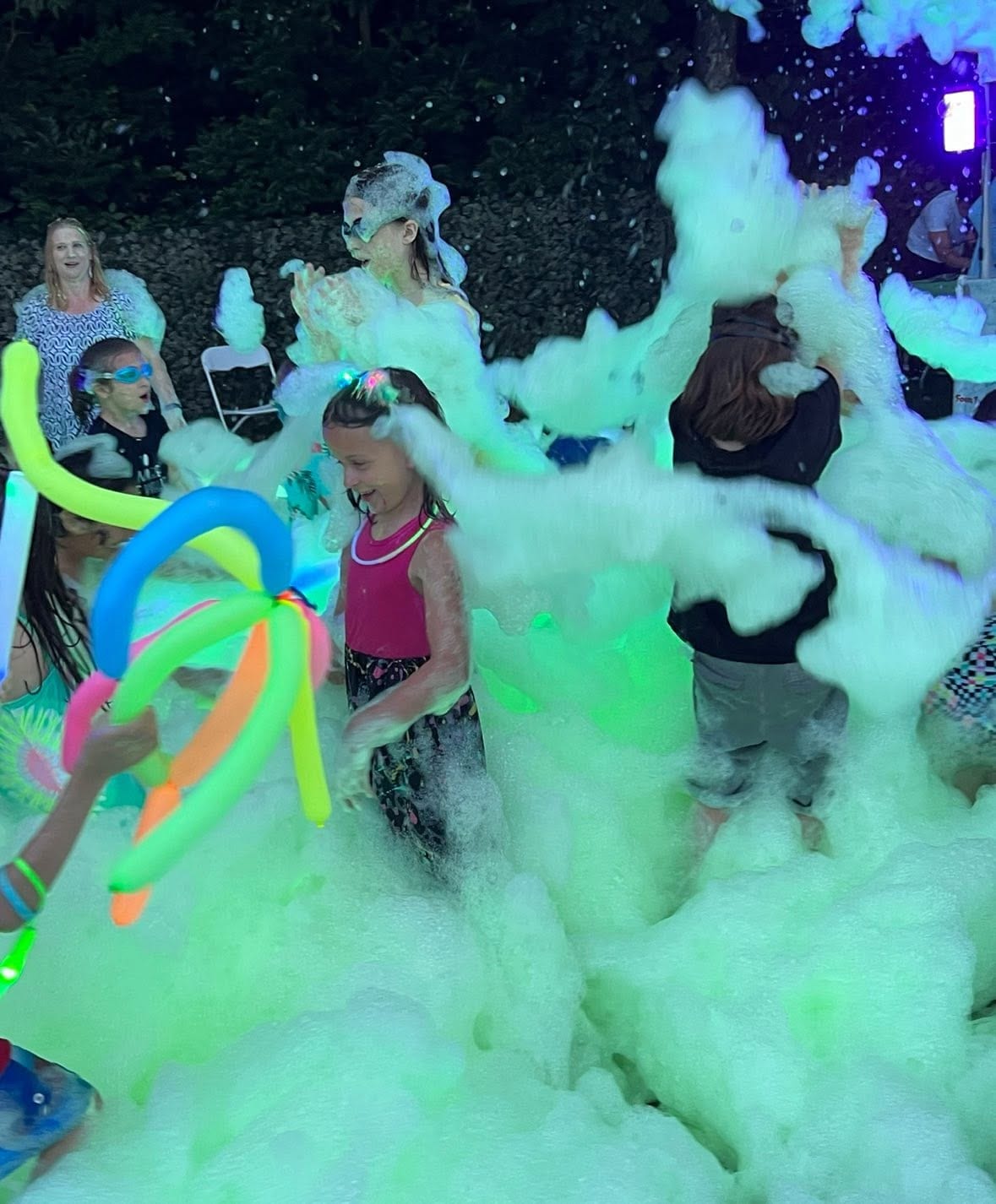 Children playing with glowing balloon swords in foam under colored light at night