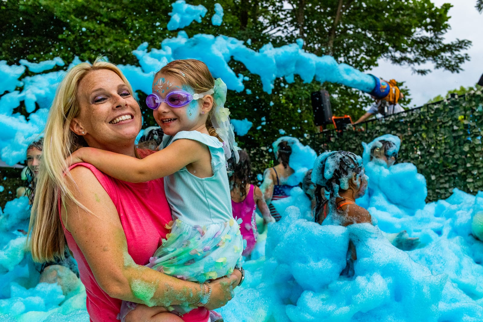 An adult holding a laughing child in a pink foam party on a lawn