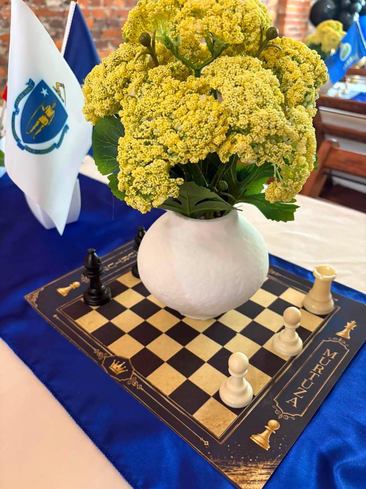 Chessboard table runner with a white vase of yellow blooms and chess pieces at a formal event table