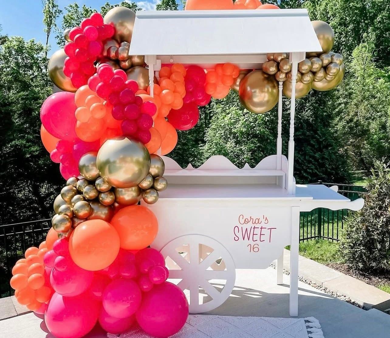 White candy cart with a hot-pink, orange and gold balloon garland for a Sweet 16 party