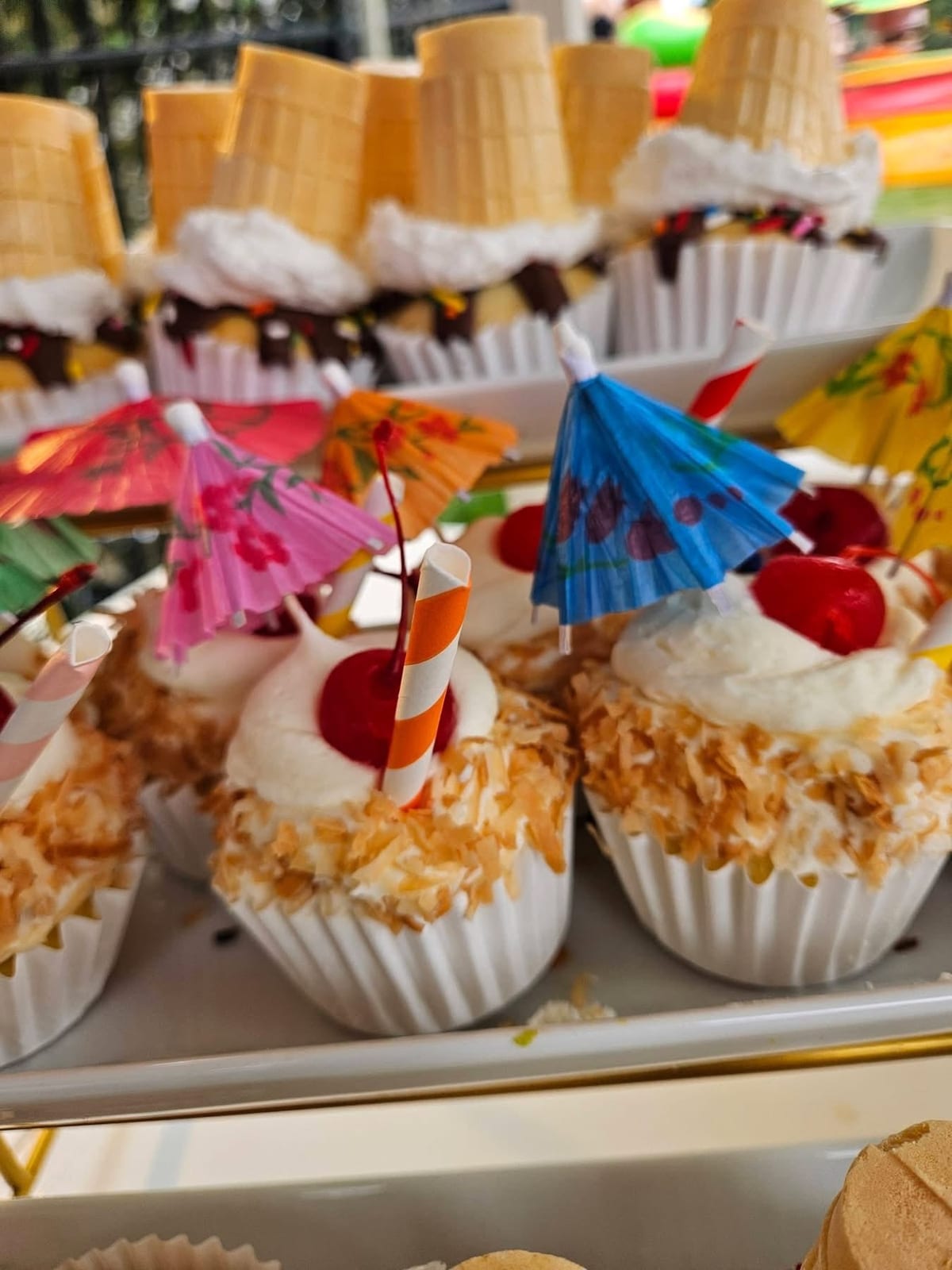 Dessert cups topped with whipped cream, cherries and paper cocktail umbrellas beside waffle cones