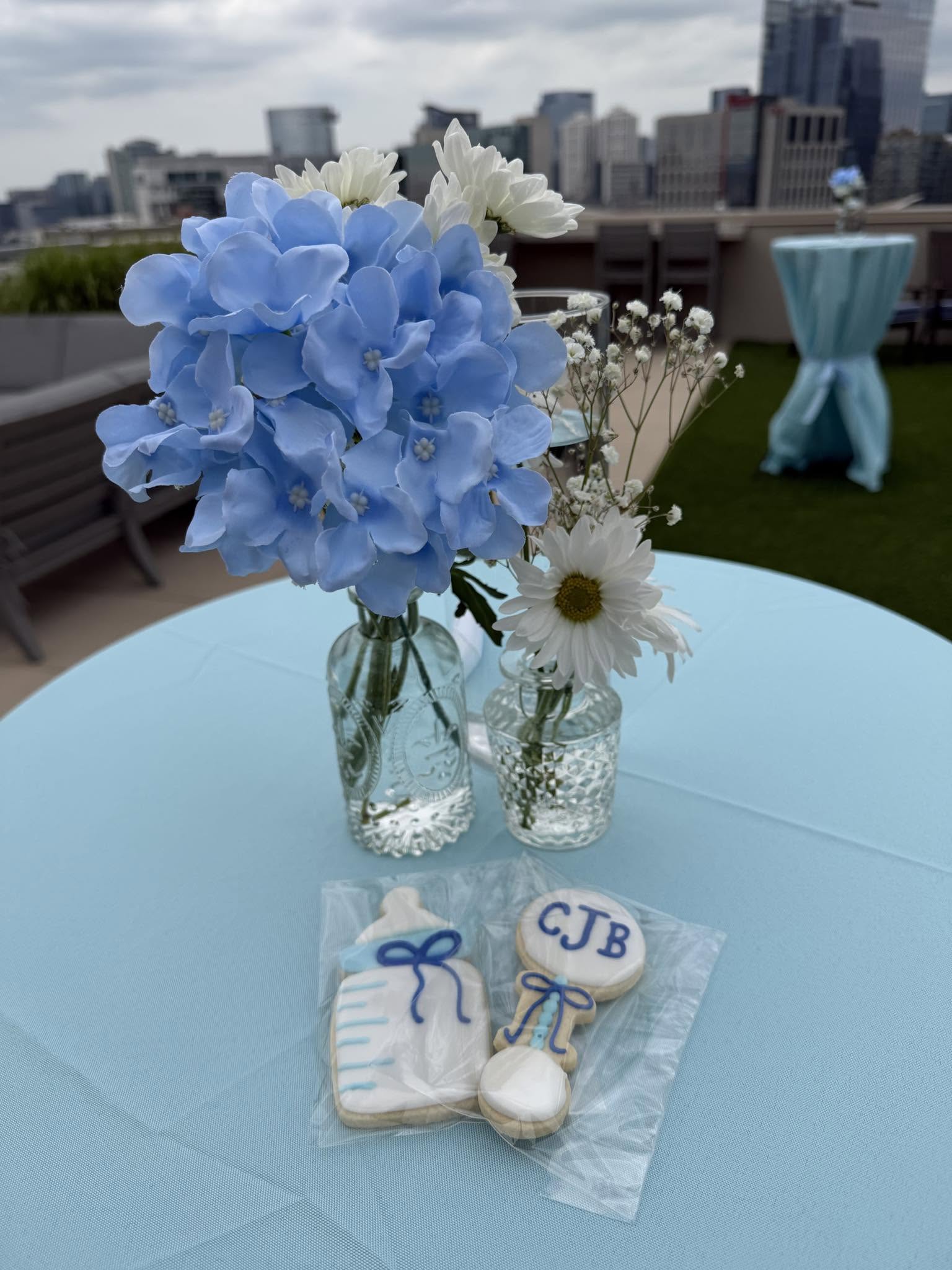 Blue hydrangeas and white daisies in cut-glass vases beside monogrammed iced cookies on a pale blue cloth, with the Nashville skyline behind