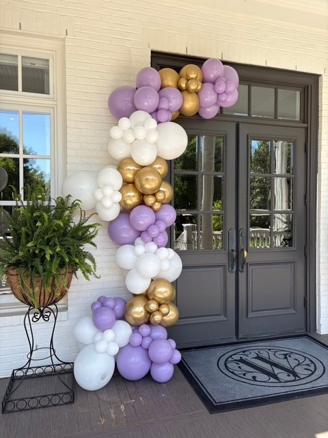 Lilac, white and chrome-gold balloon garland framing the dark double front doors of a white brick house