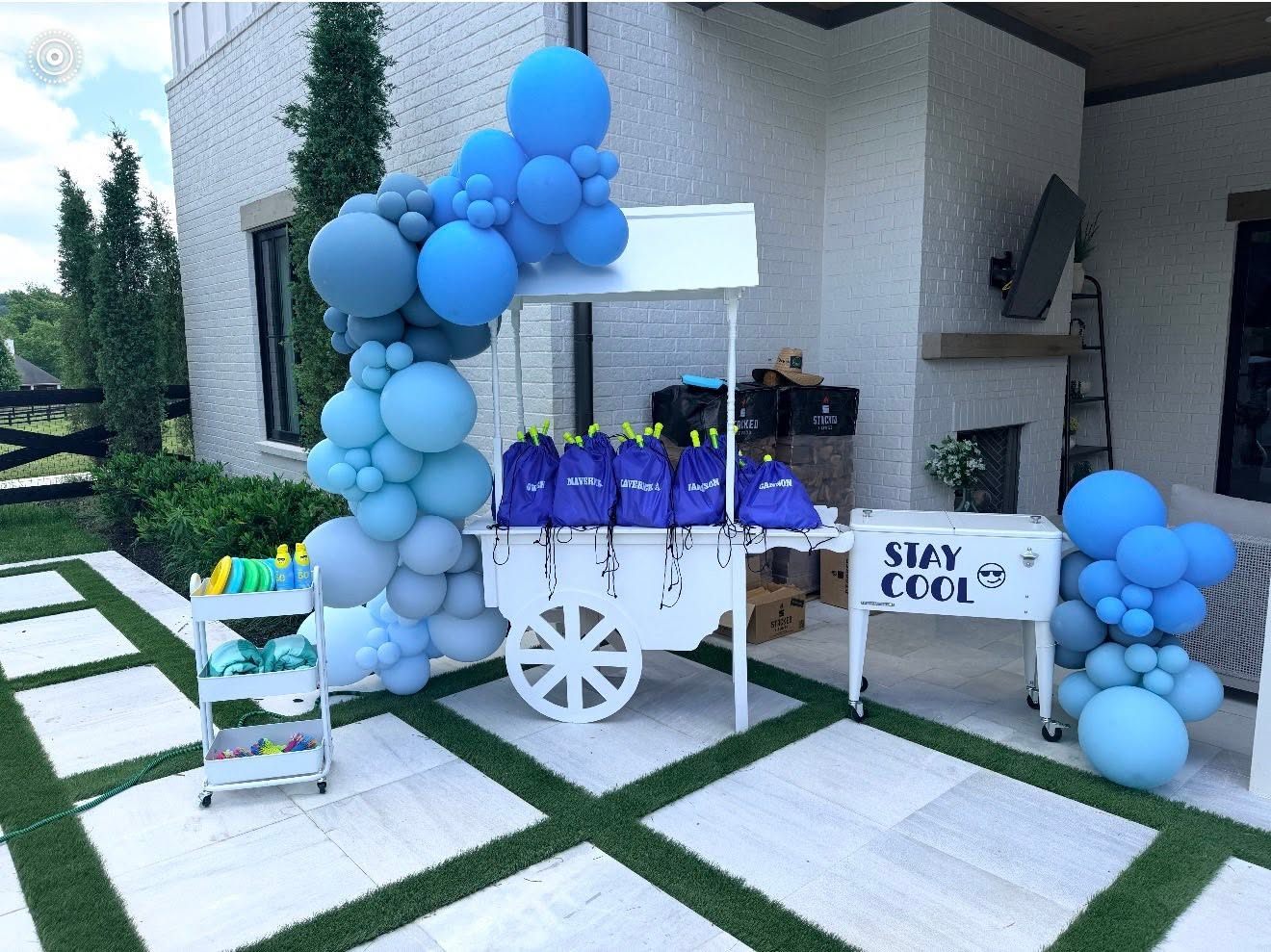 Blue balloon-arched 'Stay Cool' grad favor cart with water balloons and drawstring bags on a patio
