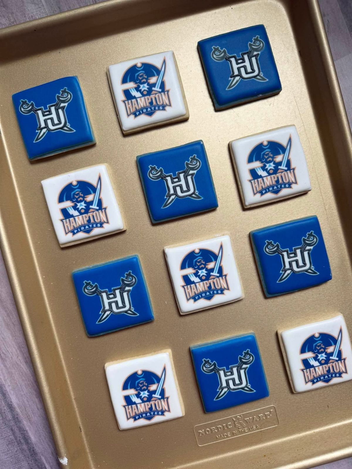 A dozen Hampton Pirates HU logo cookies in blue and white arranged on a gold baking tray