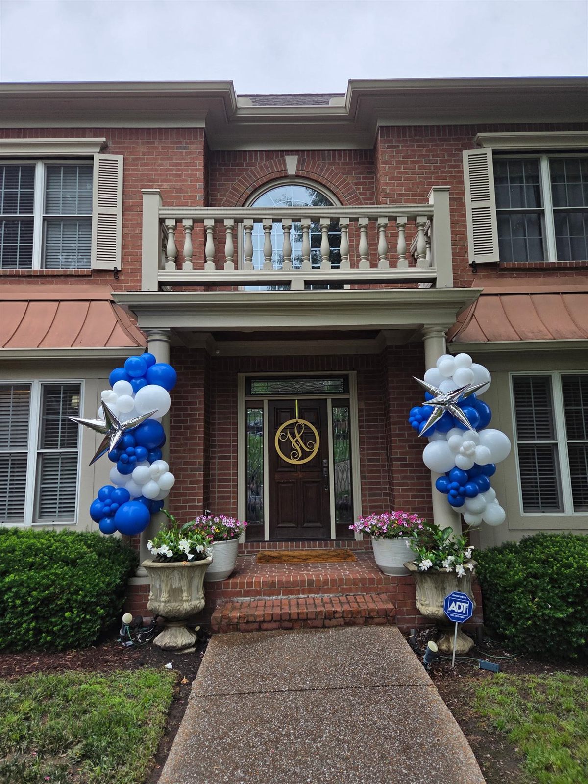 Blue, white and silver-star balloon columns framing a brick front door with a gold monogram wreath