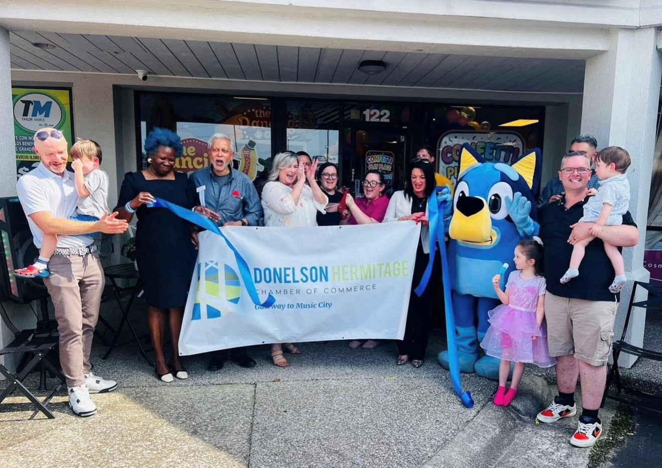 Guests holding a chamber of commerce banner and a wide blue ribbon outside a new storefront, with a costumed mascot alongside them