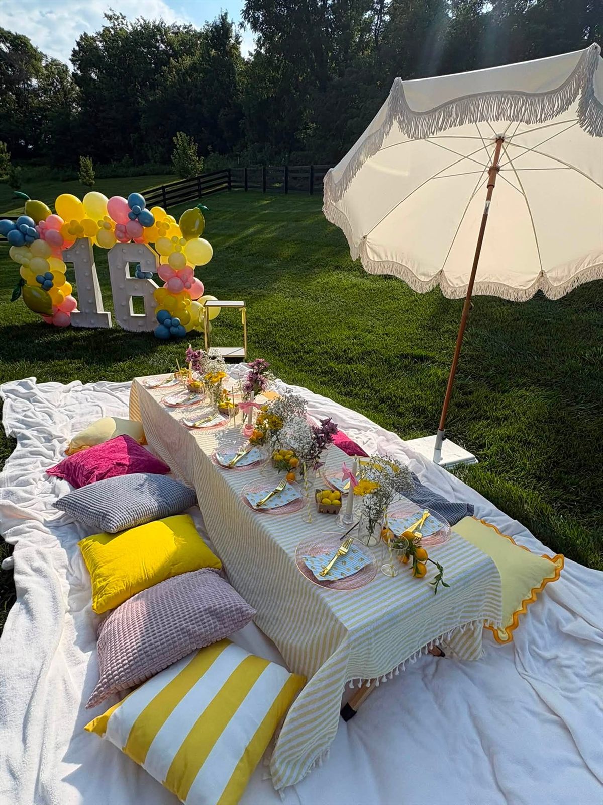 A low picnic table set with a lemon tablescape on layered blankets with colorful cushions under a fringed umbrella, with a balloon-wrapped 16 marquee behind