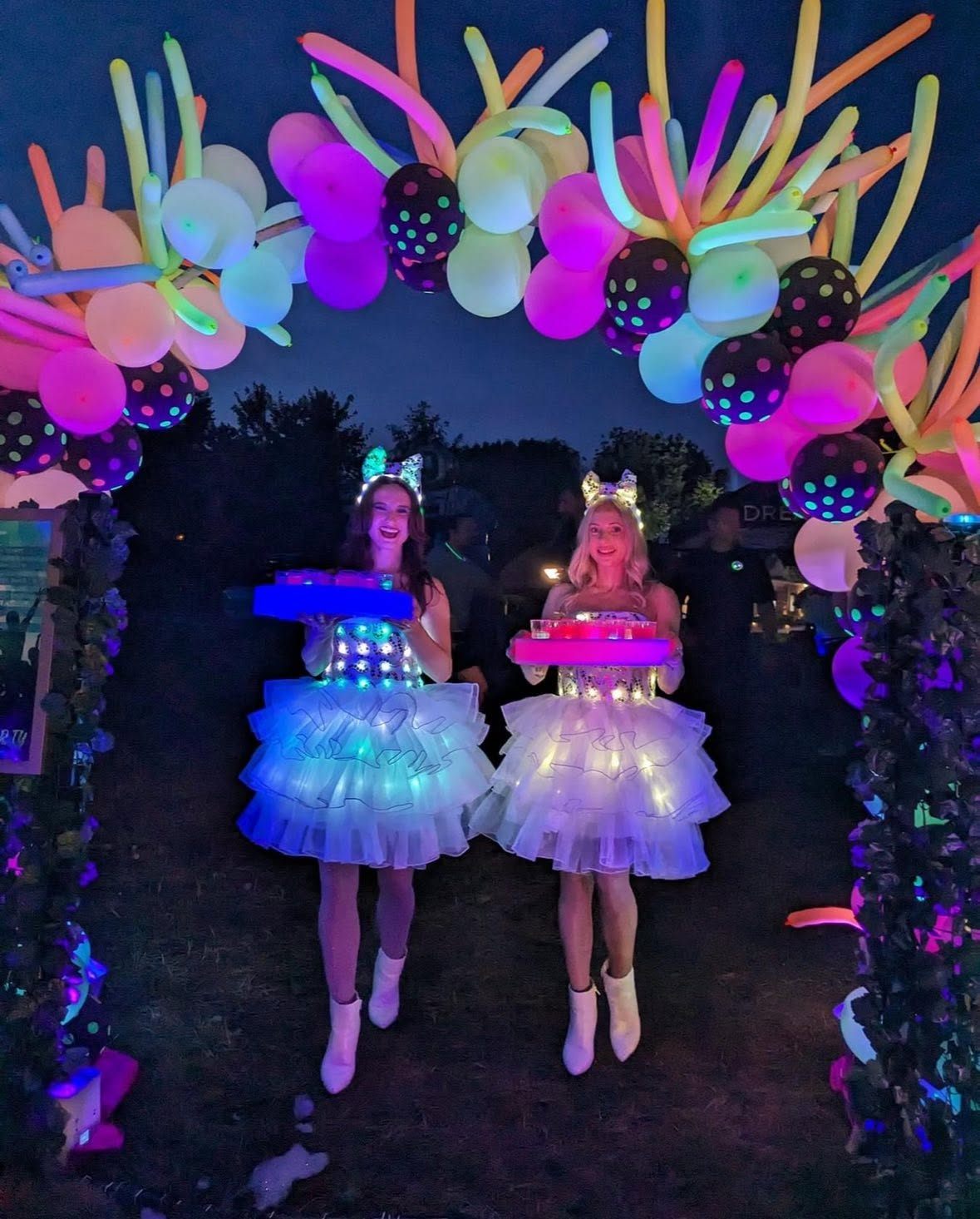 Two performers in glowing LED tutu costumes serve drinks under a neon balloon arch at night