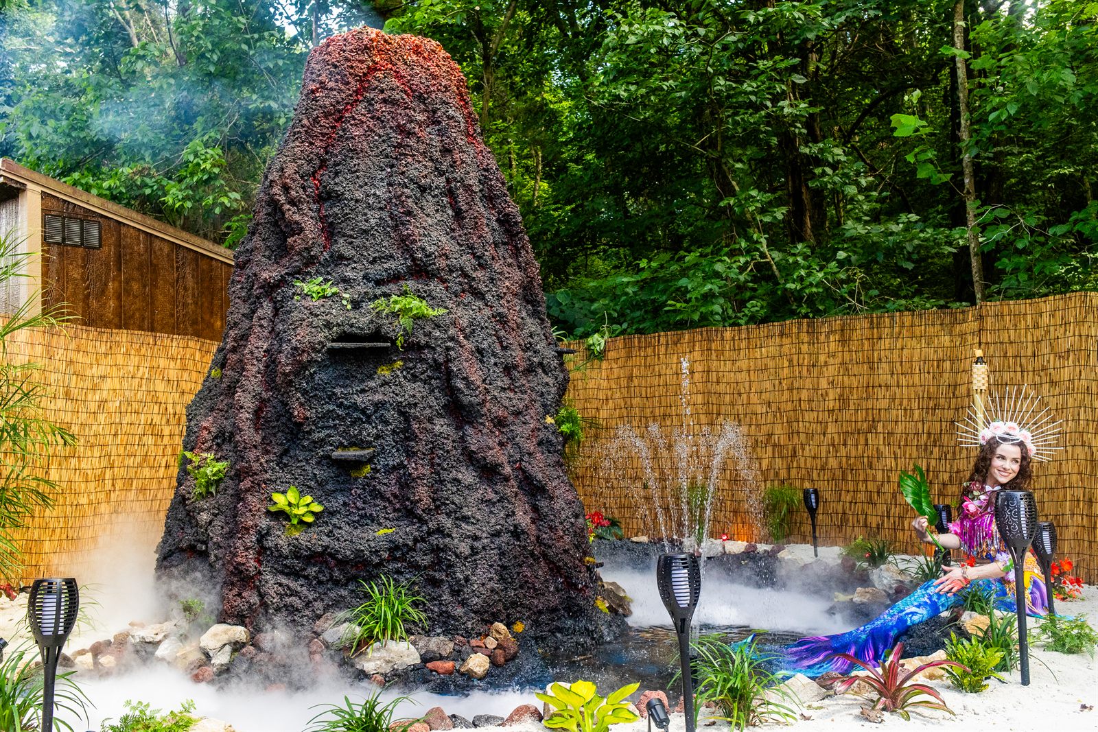 A costumed performer stands beside a smoking faux volcano with misting fountains in a garden