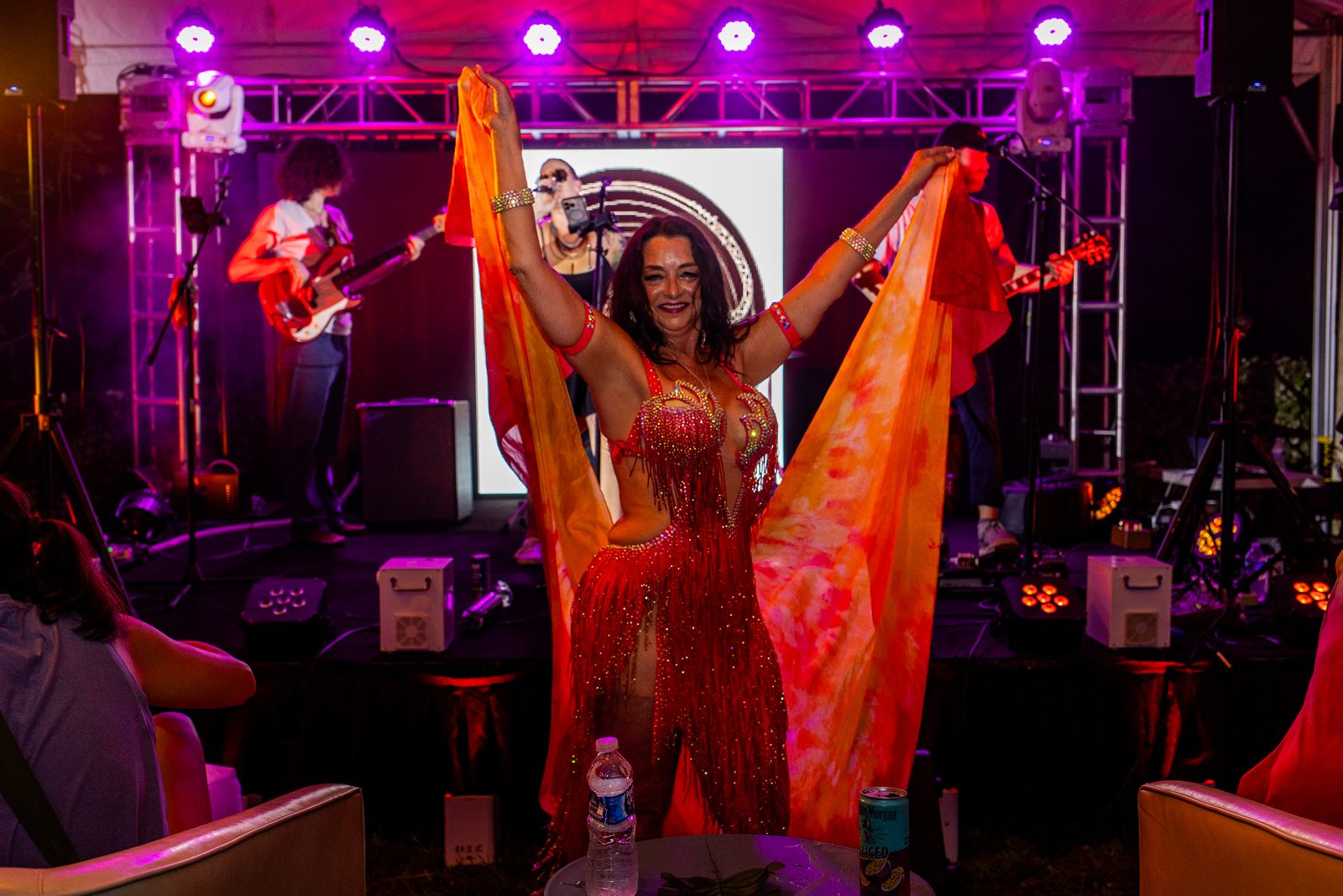 Belly dancer in fiery red-orange fringe sweeps silk veils overhead as a live band plays in magenta light