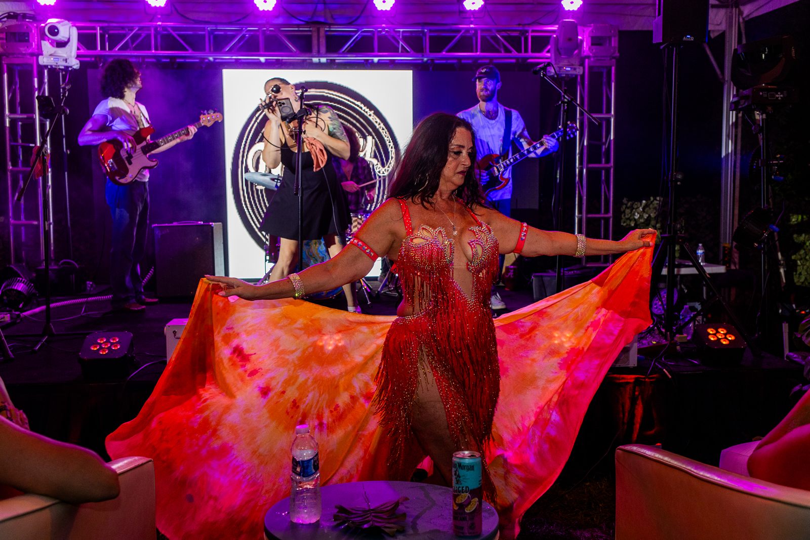 Dancer in scarlet fringe fans a tie-dyed orange silk skirt wide before a live band under purple lighting