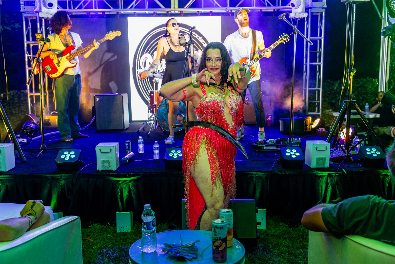 Dancer in red fringe wields a curved sword in a balance act before a live band in blue and purple light
