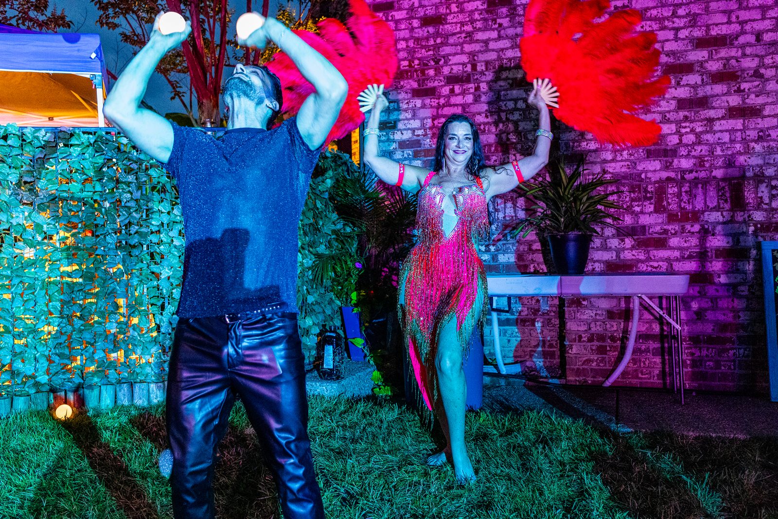 Juggler in leather tosses glowing balls beside a dancer with red feather fans against a magenta-lit wall