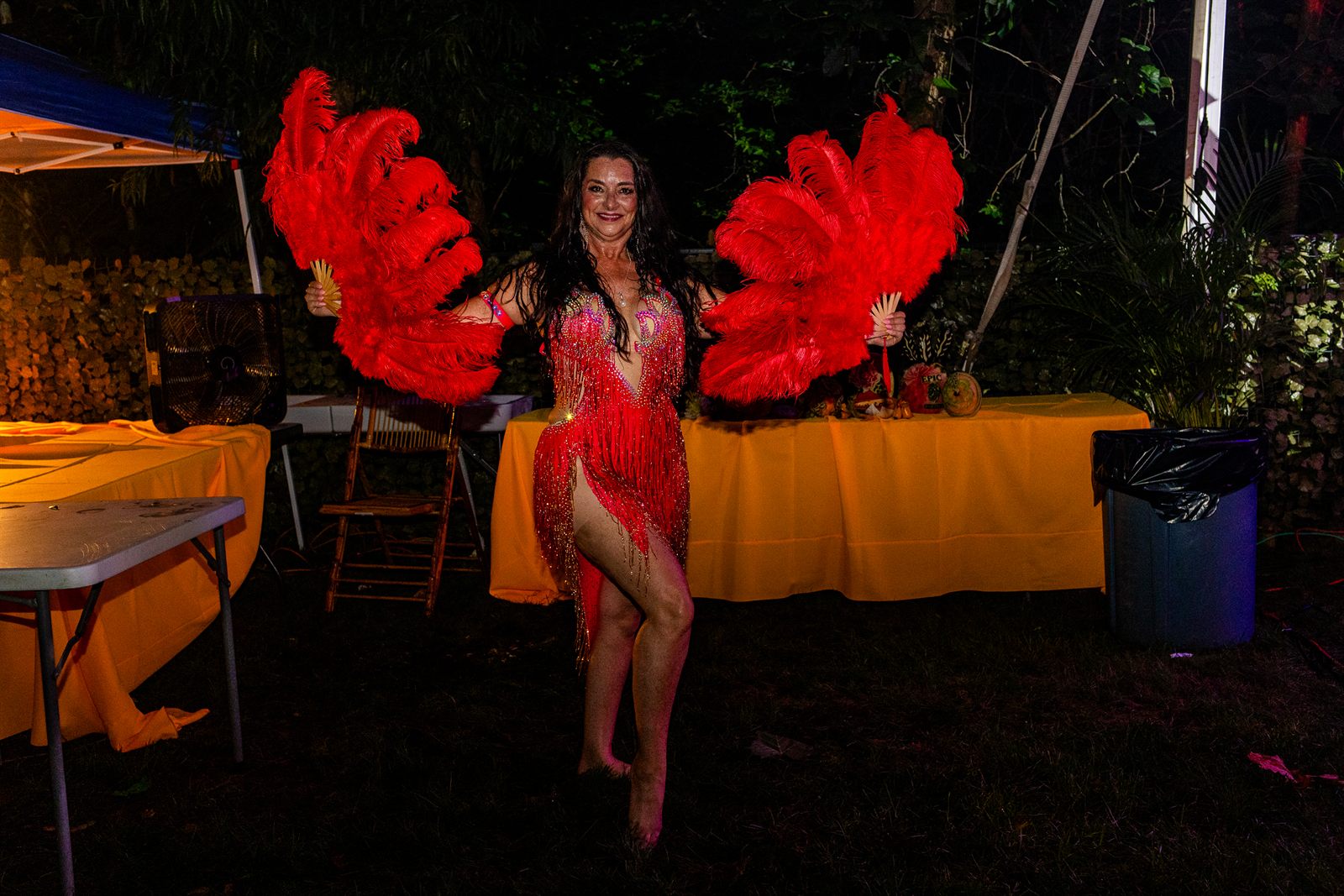 Showgirl in red sequins poses with two large red ostrich feather fans against a dark garden backdrop