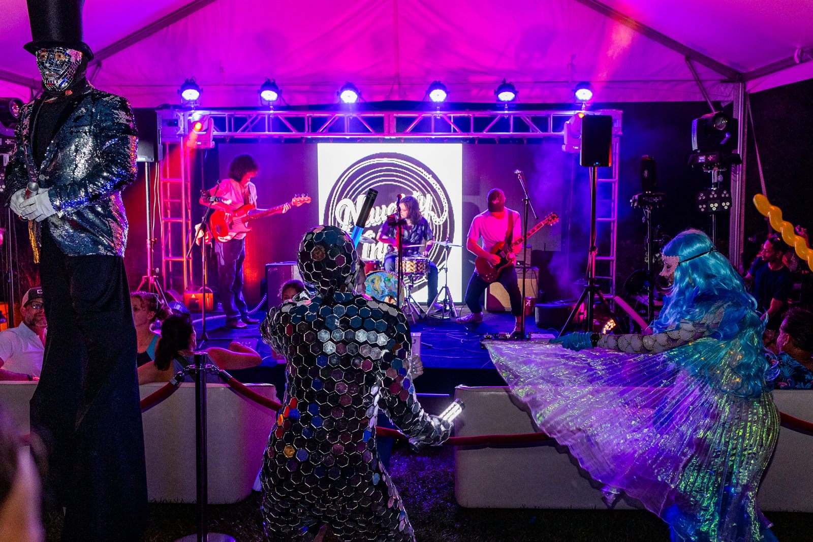 Live band on a purple-lit tent stage with a mirror-mosaic stilt walker and a shimmering LED dancer