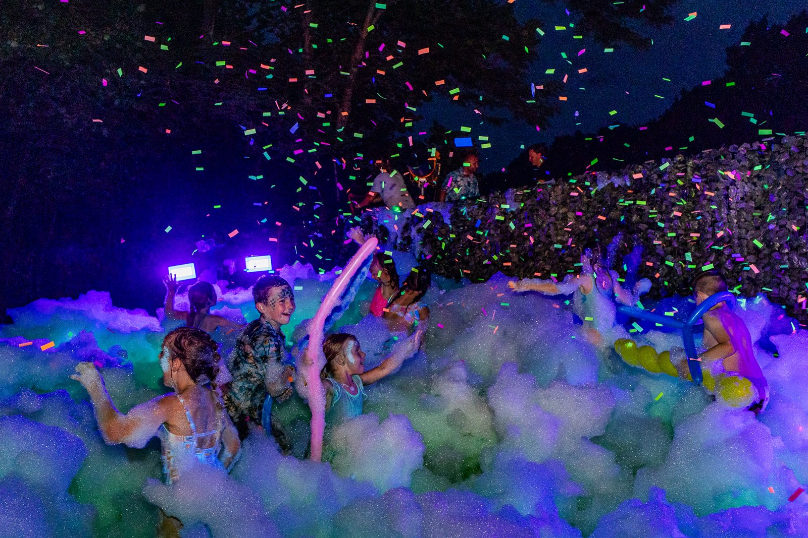 Guests in a glowing foam party under blacklight with neon confetti raining down at night