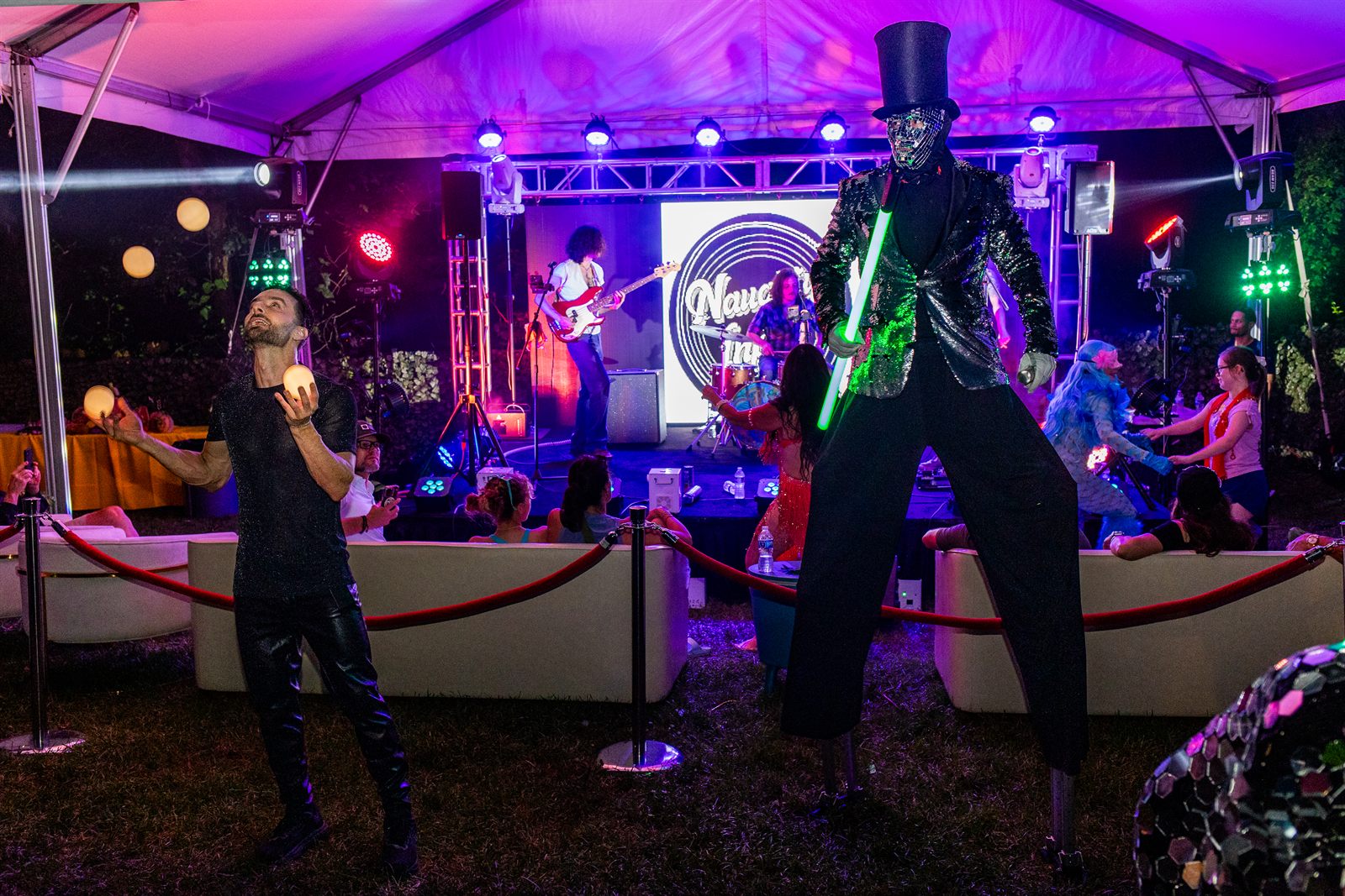 Juggler with glowing balls beside a top-hatted mirror-suited stilt walker at a purple-lit tent stage