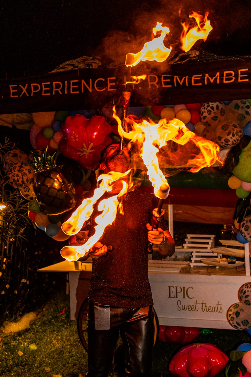 Fire performer breathing a towering plume of flame beneath an 'Experience It' balloon-arch dessert booth