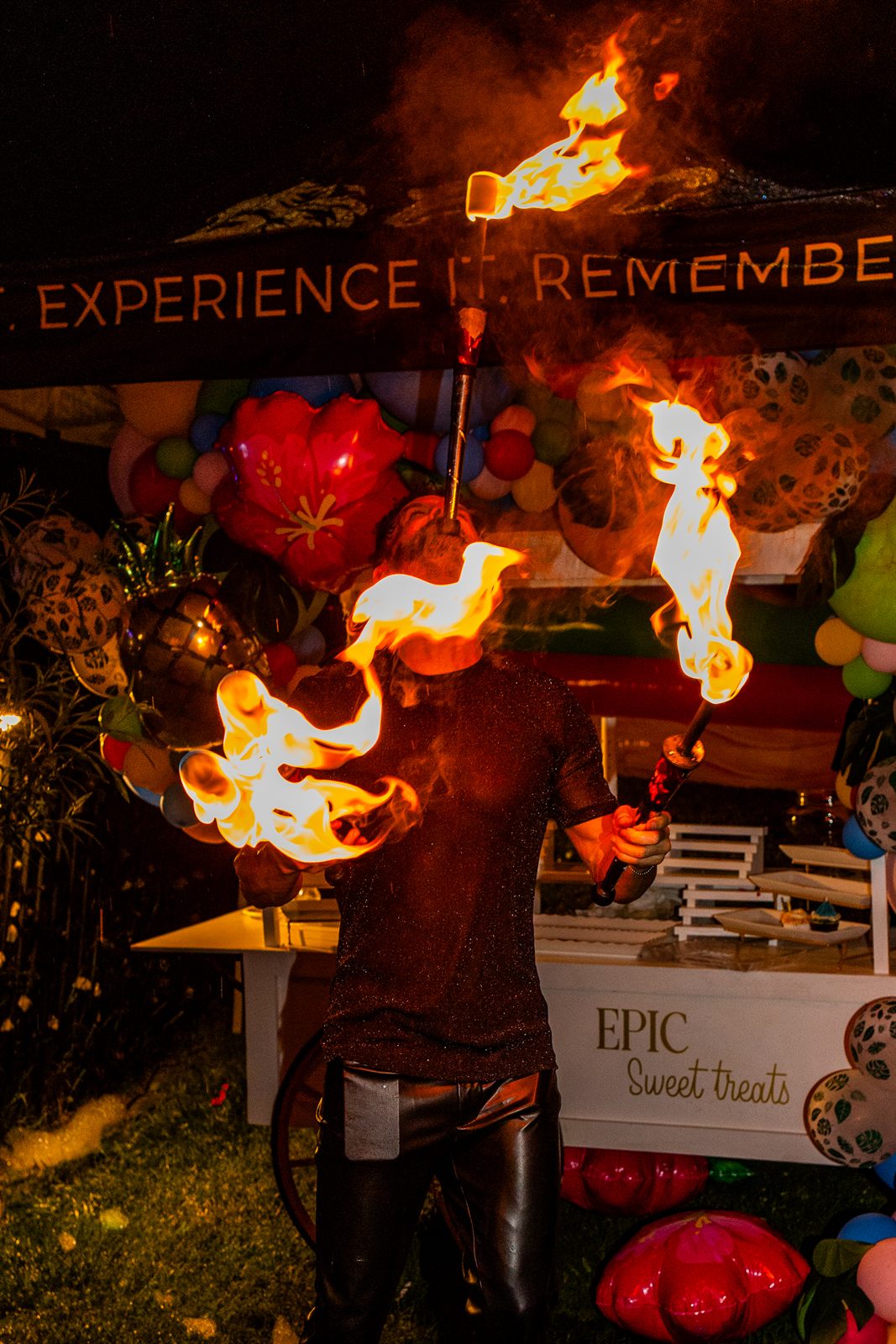 Fire eater exhaling twin bursts of flame from lit torches at a balloon-draped sweet-treats booth