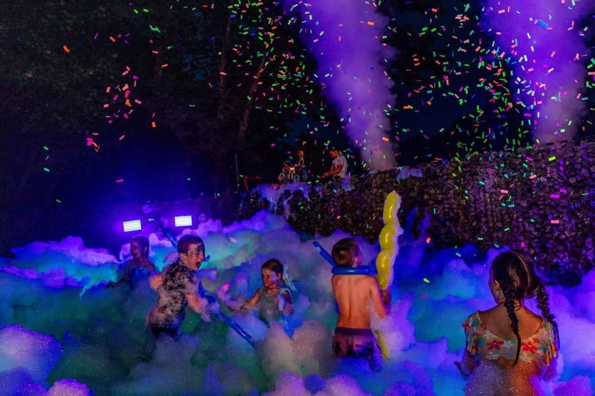 Guests dancing in glowing foam under purple blacklight as confetti falls at night
