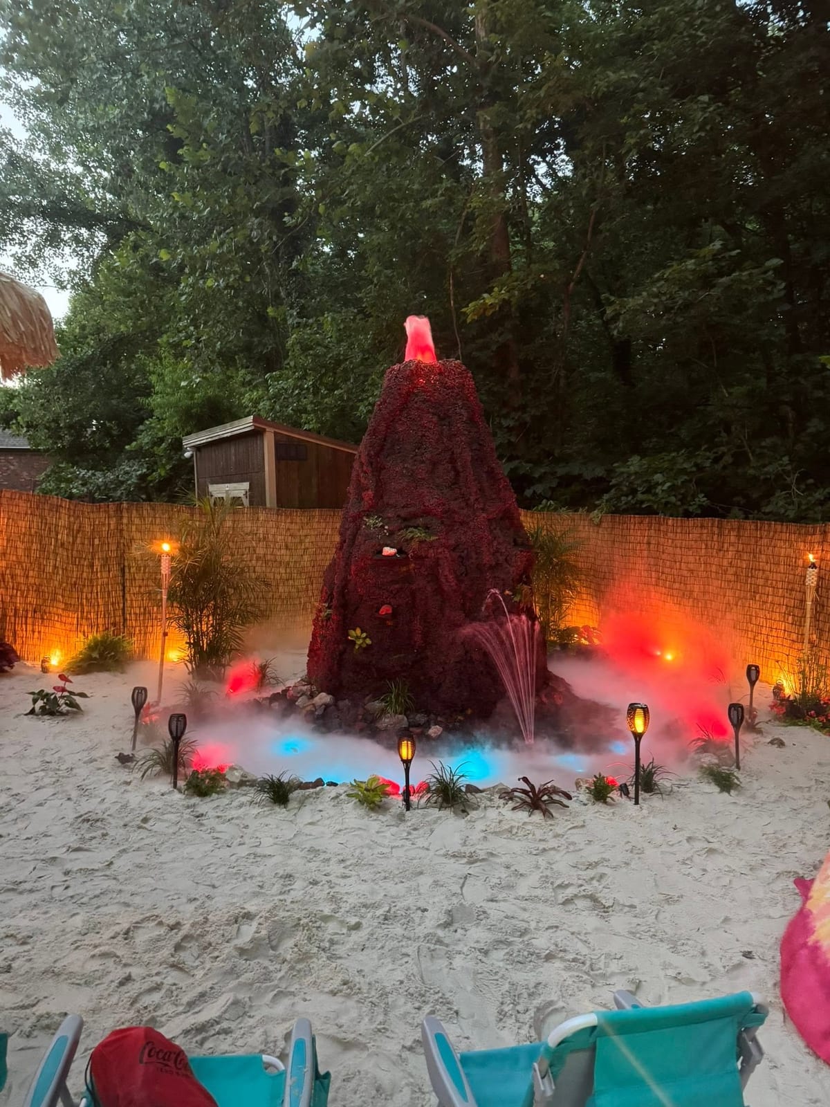 Erupting volcano centrepiece glowing red with mist over a sand beach lit by torches at dusk
