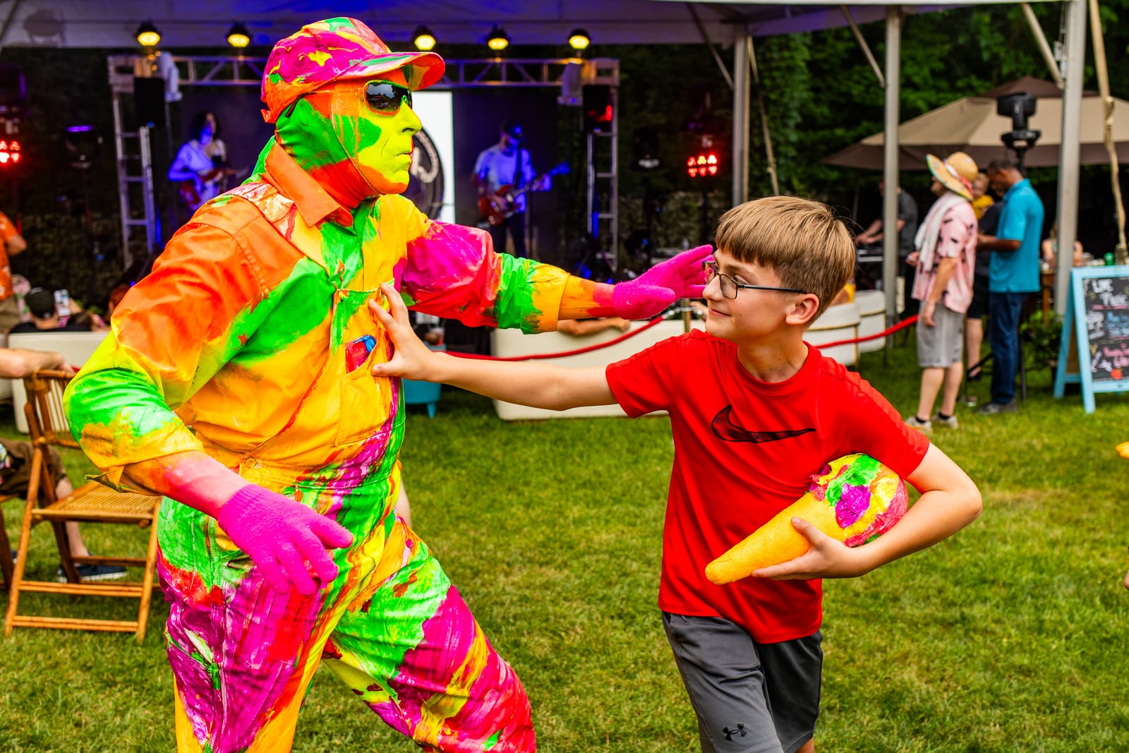 Performer in a neon rainbow suit playing a game with a young guest as a live band plays behind