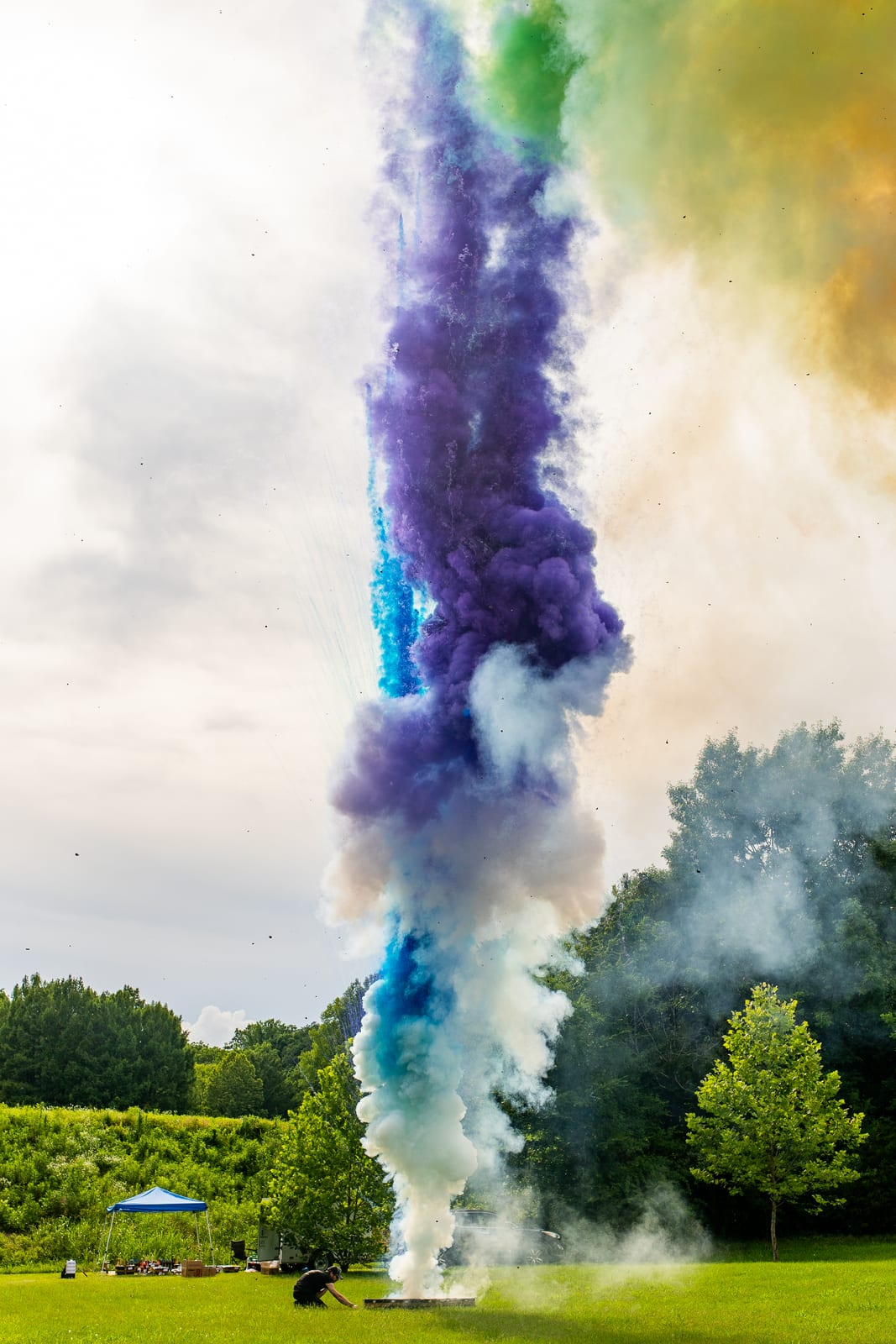 Tall column of purple, blue and green coloured smoke rising above trees on a lawn