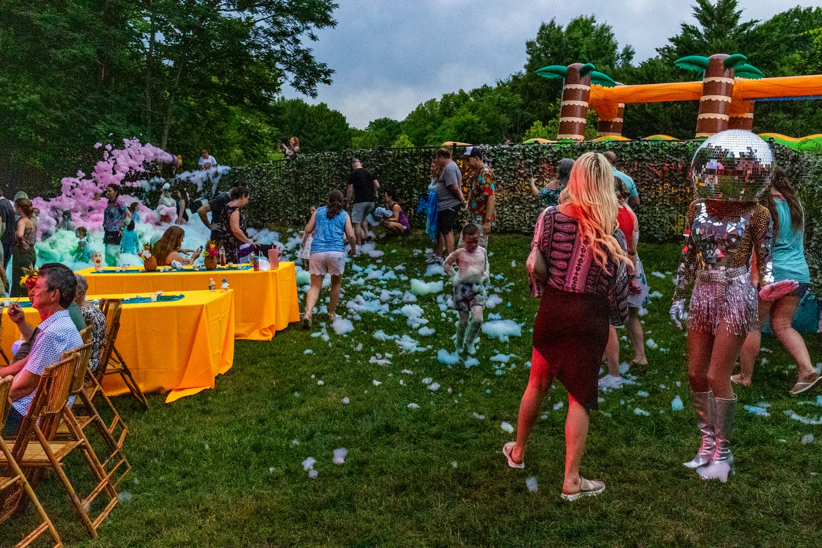 Wide view of a foam party at dusk with guests, orange banquet tables and an inflatable arch