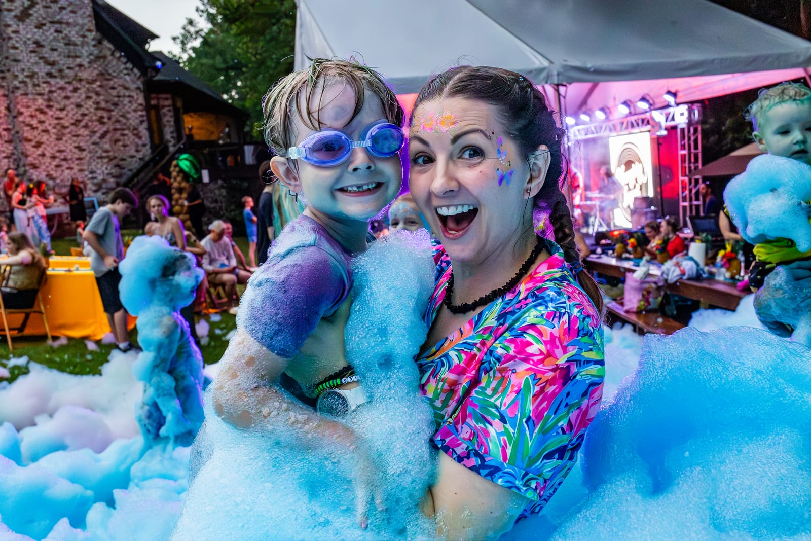 An adult and child laughing together in deep blue foam beneath colored stage lights