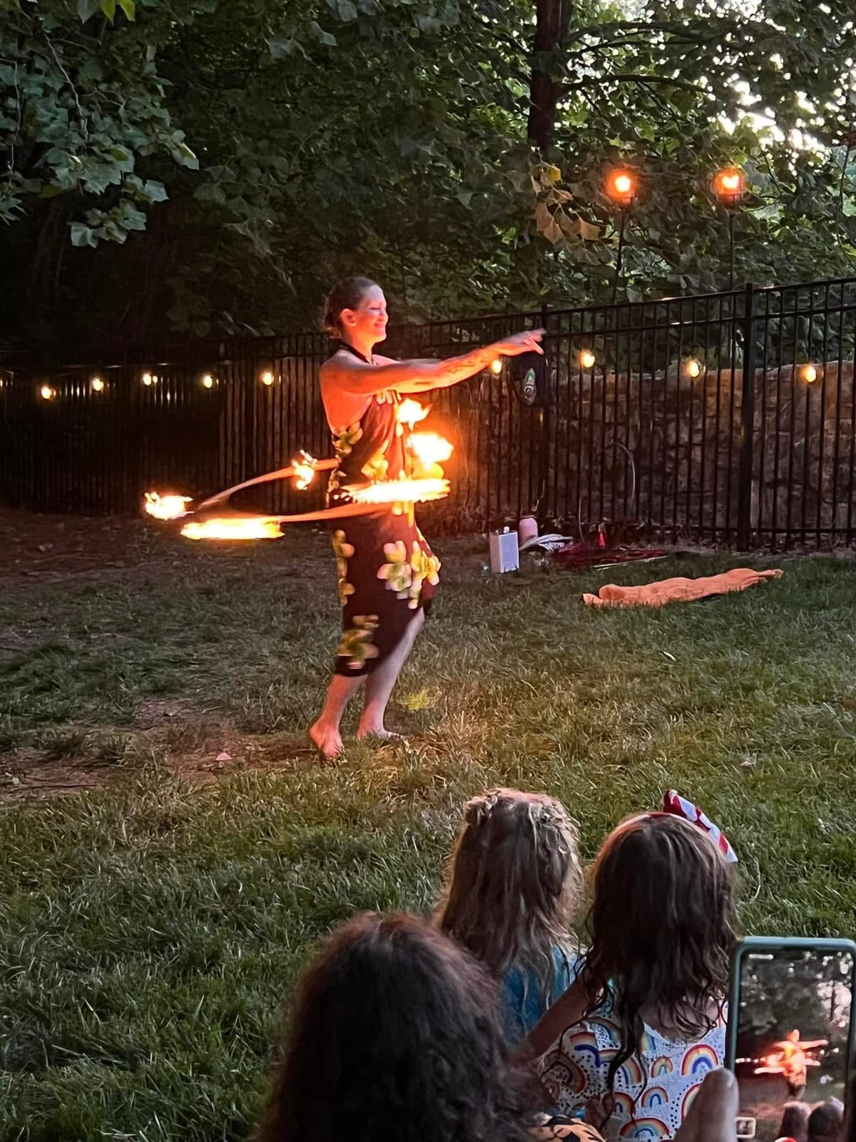 Performer spinning a flaming hoop on a lawn at dusk as guests watch