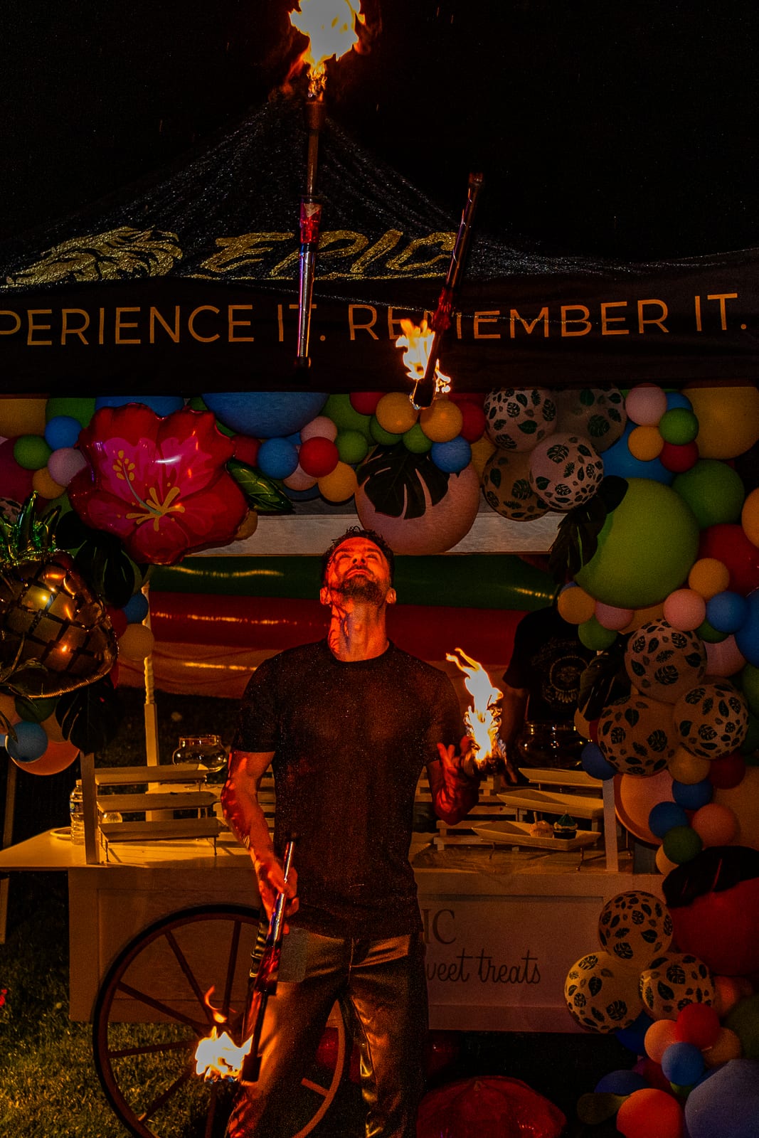 Fire performer holding lit torches beside a balloon-framed EPIC sweet cart at night